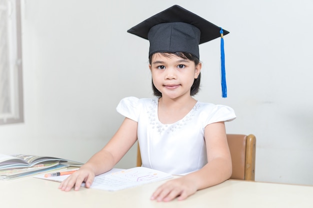 Portrait d'une petite étudiante asiatique joyeuse portant un mortier ou un chapeau de graduation tenir un crayon et écrire sur un ordinateur portable avec un fond blanc avec un espace de copie. Éducation, remise des diplômes, concept Banque de Photo