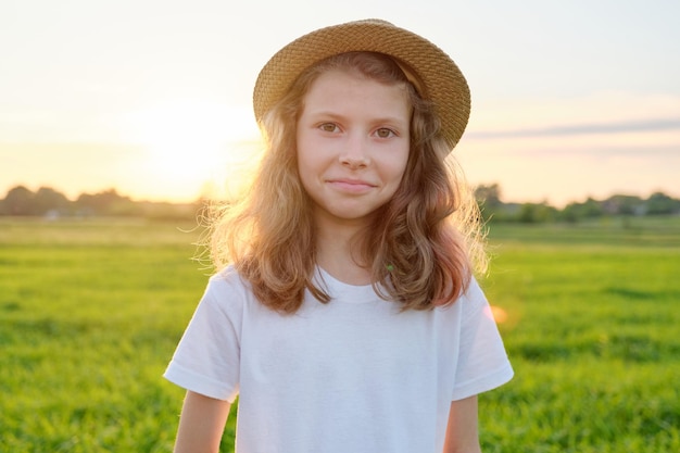 Portrait petite fille 9, 10 ans au chapeau, fond de coucher de soleil de pré vert d'été.