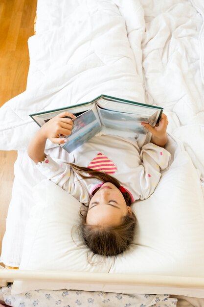 Portrait de petite fille allongée au lit le matin et lisant un gros livre