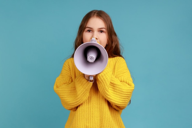 Le portrait d'une petite fille annonce des informations importantes sur l'école, regarde la caméra avec une expression calme, portant un pull jaune de style décontracté. Studio intérieur tourné isolé sur fond bleu.