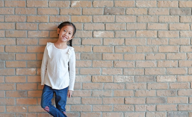 Portrait d'une petite fille asiatique heureuse avec des cheveux en queue de cochon en jeans et une chemise blanche debout au fond de mur de brique avec espace de copie.