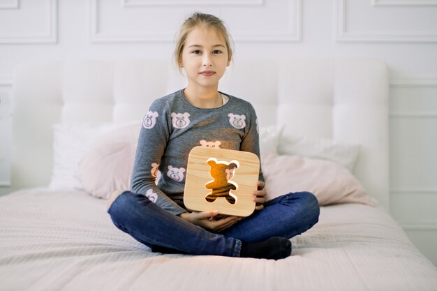 Portrait de petite fille assise sur le lit en position du lotus et tenant une lampe de nuit en bois élégante avec photo d'ours.