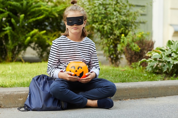 Portrait de petite fille au masque de super-héros tenant citrouille jouet dans ses mains et assis à l'extérieur