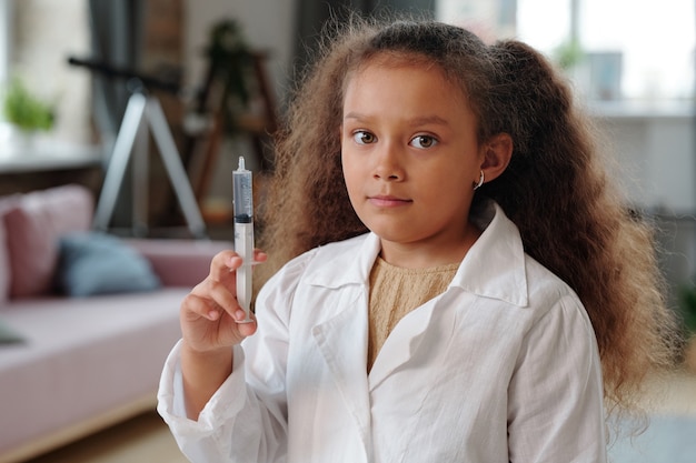 Portrait de petite fille aux cheveux bouclés et en blouse blanche tenant une seringue pendant son jeu chez le médecin