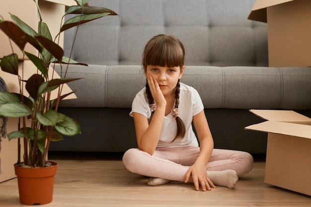 Portrait d'une petite fille aux cheveux noirs portant un t-shirt blanc assise sur le sol près d'un canapé et de boîtes en carton regardant la caméra tenant la main sous le menton déménageant