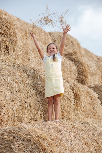 Portrait d'une petite fille bienheureuse souriante avec des dents debout sur une haute botte de foin vomissant du foin et le regardant portant une robe d'été S'amuser loin de la ville sur un champ plein de foin doré