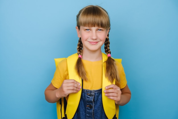 Photo portrait d'une petite fille blonde caucasienne souriante, écolière enfant tenant un sac à dos jaune debout isolée sur fond de couleur bleue en studio, regardant la caméra de manière positive. retour au concept de l'école