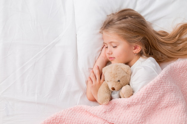 portrait d'une petite fille blonde dormant dans un lit avec un ours en peluche sous une couverture rose