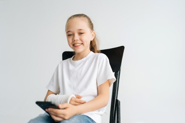 Portrait d'une petite fille blonde positive avec un bras cassé enveloppé dans un bandage en plâtre blanc souriant regardant la caméra tenant un téléphone portable assis sur une chaise