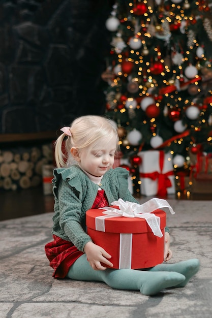 Portrait d'une petite fille blonde de trois ans avec un grand arbre de Noël cadeau rouge et des guirlandes d'ambiance de Noël