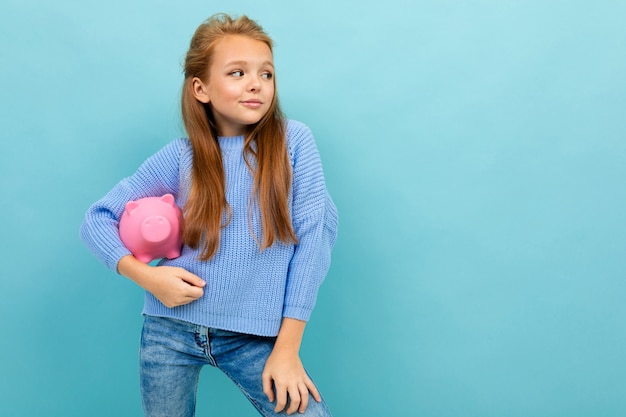 Portrait de petite fille caucasienne aux cheveux longs détient tirelire cochon rose isolé sur bleu