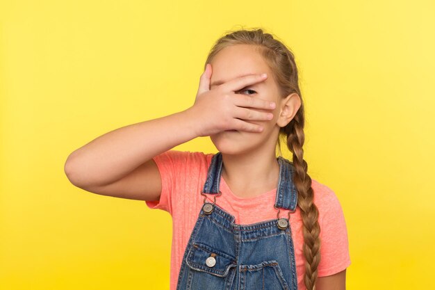 Portrait d'une petite fille curieuse avec une tresse en salopette en denim couvrant le visage avec la main et regardant à travers les doigts enfant lorgnant timidement sur un plan secret en studio isolé sur fond jaune