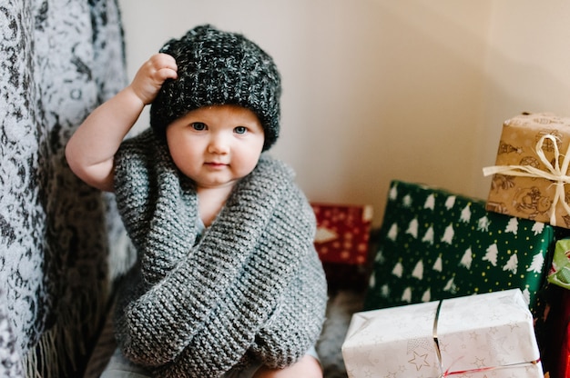 Portrait d'une petite fille dans une écharpe tricotée chaude, enlève le chapeau, s'assoit dans la chambre avec des cadeaux.