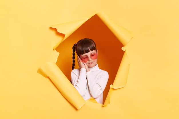 Portrait d'une petite fille endormie et fatiguée avec des tresses et des lunettes portant une chemise décontractée regardant à travers un trou déchiré dans du papier jaune garde les paumes près des joues et les yeux fermés