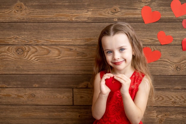 Portrait d'une petite fille enfant vêtue d'une robe rouge tenant un jouet coeur rouge sur un fond en bois brun foncé et souriant doucement le concept de la Saint-Valentin un espace vide pour le texte