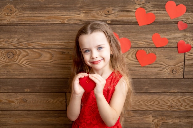 Portrait d'une petite fille enfant vêtue d'une robe rouge tenant un jouet coeur rouge sur un fond en bois brun foncé et souriant doucement le concept de la Saint-Valentin un espace vide pour le texte
