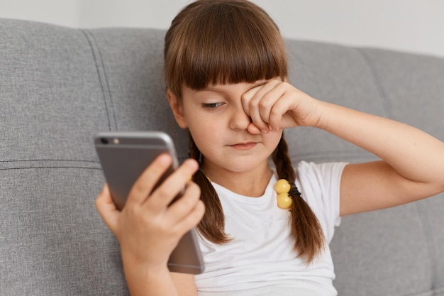 Portrait d'une petite fille épuisée portant un t-shirt blanc assise sur un canapé à la maison avec un téléphone portable, jouant à des jeux ou regardant des dessins animés longtemps, se frottant les yeux, a l'air endormi et fatigué.