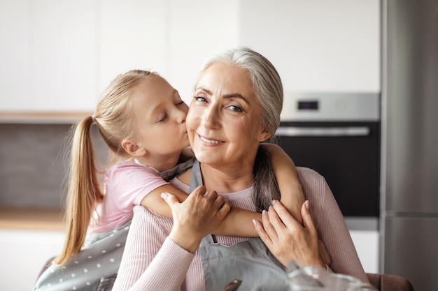 Photo portrait d'une petite-fille européenne souriante embrasse la joue d'une grand-mère âgée en tablier