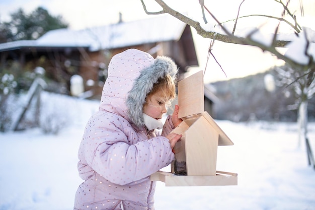 Portrait de petite fille à l'extérieur dans le jardin d'hiver, debout près de la mangeoire à oiseaux en bois.