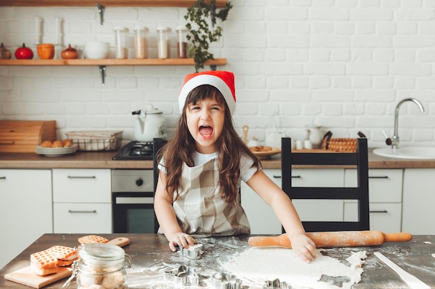 Portrait d'une petite fille heureuse dans un bonnet de Noel roulant la pâte sur la table de la cuisine un enfant préparant des biscuits de Noël