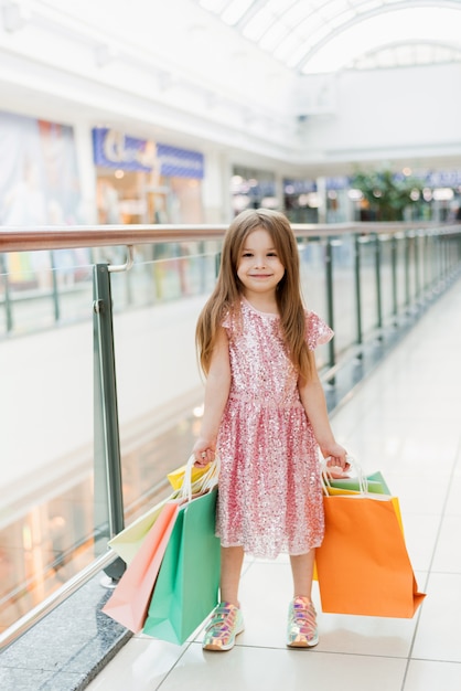 Portrait d'une petite fille heureuse dans le centre commercial. Une jeune fille riante souriante dans une robe rose avec des sacs multicolores dans ses mains est engagée dans le shopping. Modèle pour votre annonce