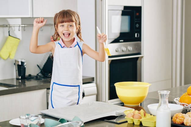 Portrait de petite fille heureuse excitée avec une brosse en silicone à la main appréciant la cuisson des biscuits à la maison