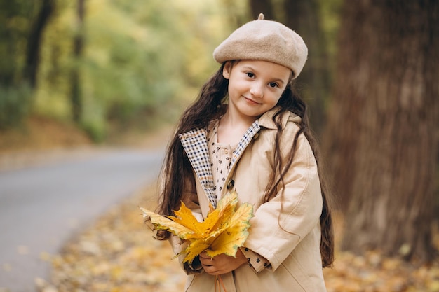 Portrait d'une petite fille heureuse en manteau beige et béret tenant des feuilles d'érable jaunes et passant du temps dans le parc d'automne