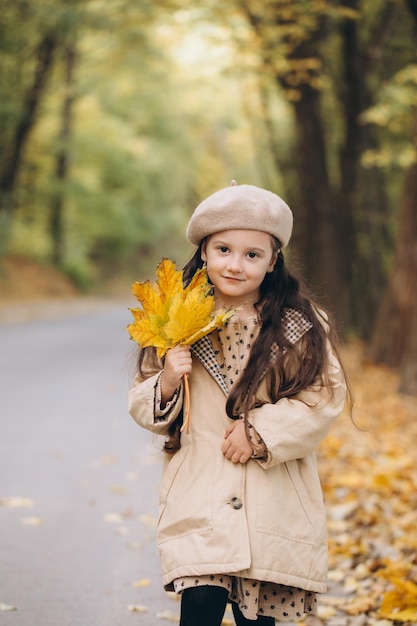 Portrait d'une petite fille heureuse en manteau beige et béret tenant des feuilles d'érable jaunes et passant du temps dans le parc d'automne