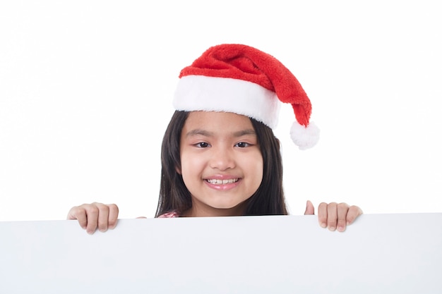 Portrait d'une petite fille heureuse portant bonnet de Noel posant derrière un panneau blanc isolé sur blanc