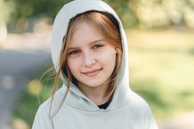 Portrait d'une petite fille heureuse en sweat à capuche souriant à la caméra en marchant dans un parc sur une nature floue ba...