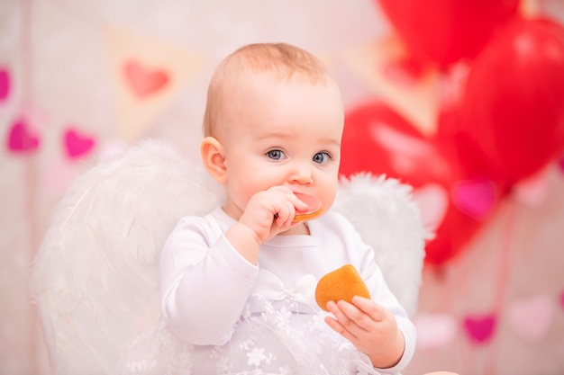 Portrait d'une petite fille joyeuse avec des ailes de plumes blanches, manger des cookies en forme de coeur