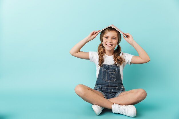 Portrait d'une petite fille joyeuse assise isolée sur un mur bleu, tenant un livre sur la tête