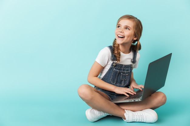 Portrait d'une petite fille joyeuse isolée sur un mur bleu, étudiant avec un ordinateur portable