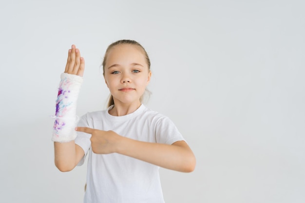 Portrait d'une petite fille joyeuse montrant avec l'index à la main cassée enveloppée dans un bandage en plâtre blanc avec un tirage coloré regardant la caméra