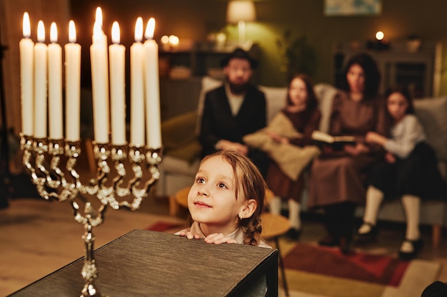 Portrait d'une petite fille juive regardant la bougie menorah avec admiration tout en célébrant hanukkah avec fami