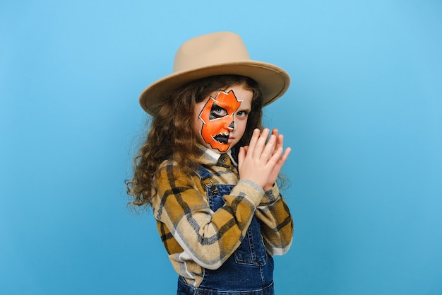 Photo portrait d'une petite fille avec un masque de maquillage porte un chapeau marron dit boo rit, effrayant souriant regardant la caméra, posant isolé sur fond bleu en studio. concept de vacances halloween heureux