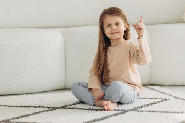 Portrait d'une petite fille mignonne Elle est assise sur le sol près du canapé La fille regarde la caméra et montre un doigt vers le haut