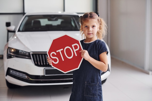 Portrait d'une petite fille mignonne qui tient un panneau de signalisation dans les mains dans un salon automobile.