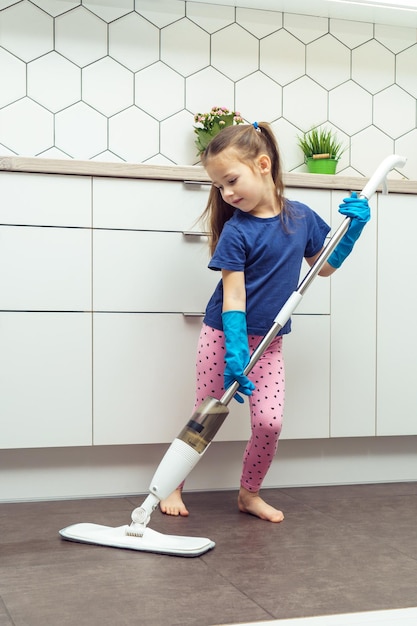 Portrait d'une petite fille préadolescente occupée portant des gants en caoutchouc bleu tenant une vadrouille à main pour laver le sol dans la cuisine