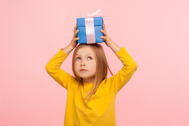 Portrait d'une petite fille réfléchie tenant un cadeau sur la tête et levant les yeux avec un visage pensif, curieuse de ce qu'il y a à l'intérieur de la boîte, rêvant d'un bon cadeau. studio d'intérieur tourné isolé sur fond rose