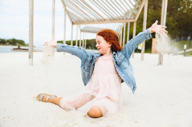 Portrait d'une petite fille rousse vêtue d'une robe assise sur le sable de la plage Journée ensoleillée d'été