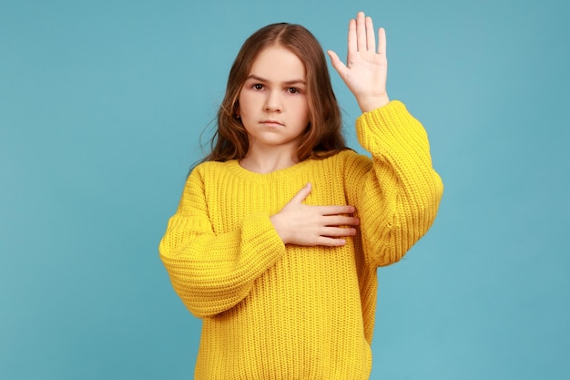 Portrait d'une petite fille sérieuse levant la paume pour prêter serment, enfant jurant de ne dire que la vérité, portant un pull jaune de style décontracté. Studio intérieur tourné isolé sur fond bleu.