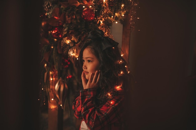 Portrait d'une petite fille souriante asiatique candide en pyjama à carreaux rouge assis sur des marches en bois à Noël