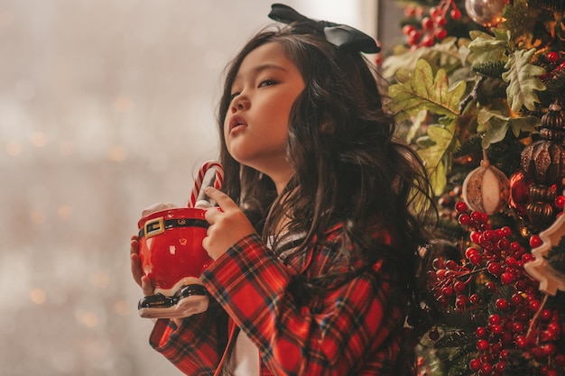 Portrait d'une petite fille souriante asiatique candide tenant une tasse de Noël rouge avec des guimauves et une canne en bonbon