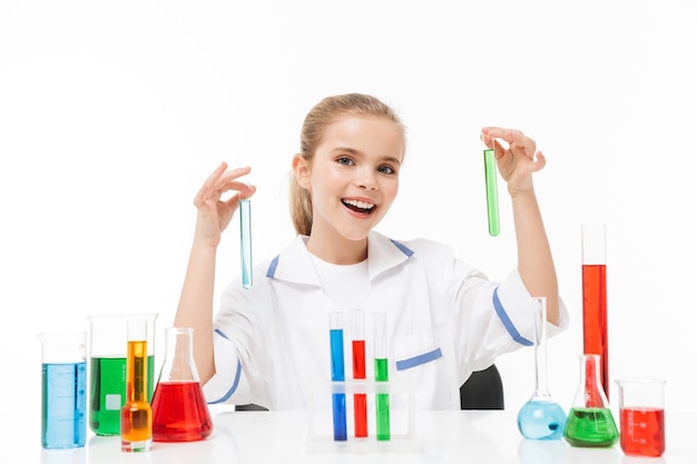 Portrait d'une petite fille souriante en blouse blanche de laboratoire faisant des expériences chimiques avec un liquide multicolore dans des tubes à essai isolés sur un mur blanc