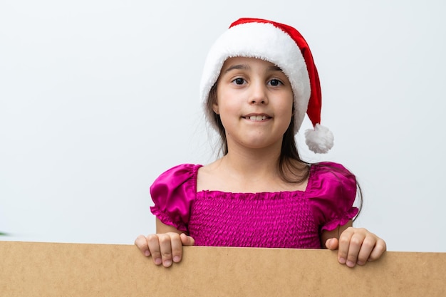 Portrait de petite fille souriante avec chapeau de Noël. Kid lorgnant derrière le tableau des vacances vierges. Heureux enfant mignon posant derrière un panneau de papier kraft, isolé sur fond blanc.