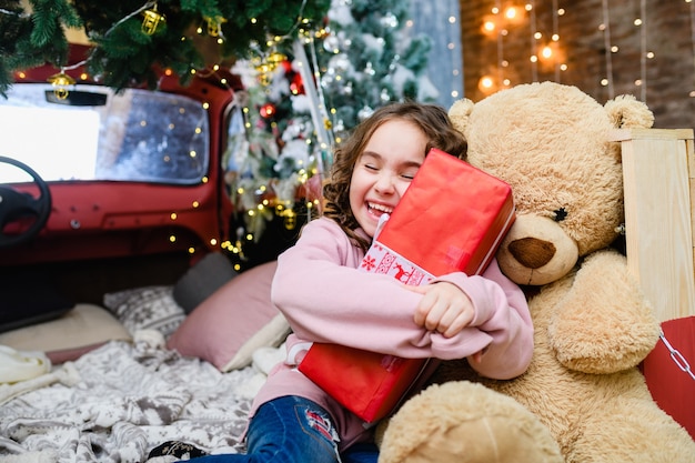 Portrait d'une petite fille souriante tenant une grande boîte cadeau rouge près d'un sapin décoré de Noël et du nouvel an, une enfance heureuse et des vacances d'hiver