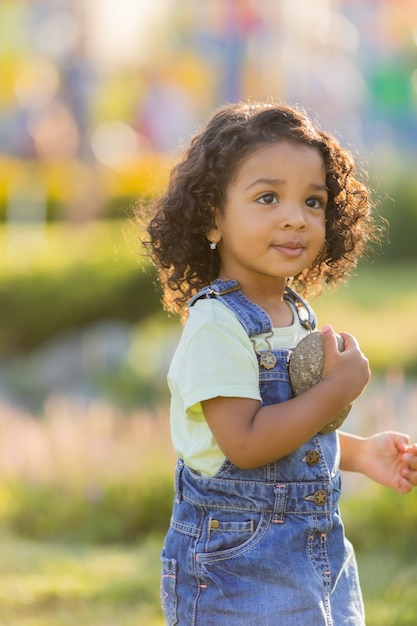 Portrait petite fille de teint basané ludique dans une robe d'été en jean debout dans le jardin aux beaux jours