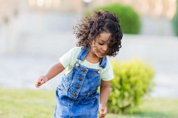 Portrait petite fille de teint basané ludique dans une robe d'été en jean debout dans le jardin aux beaux jours