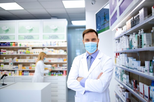 Portrait de pharmacien portant un masque et une blouse blanche debout dans un magasin de pharmacie pendant la pandémie de virus corona.